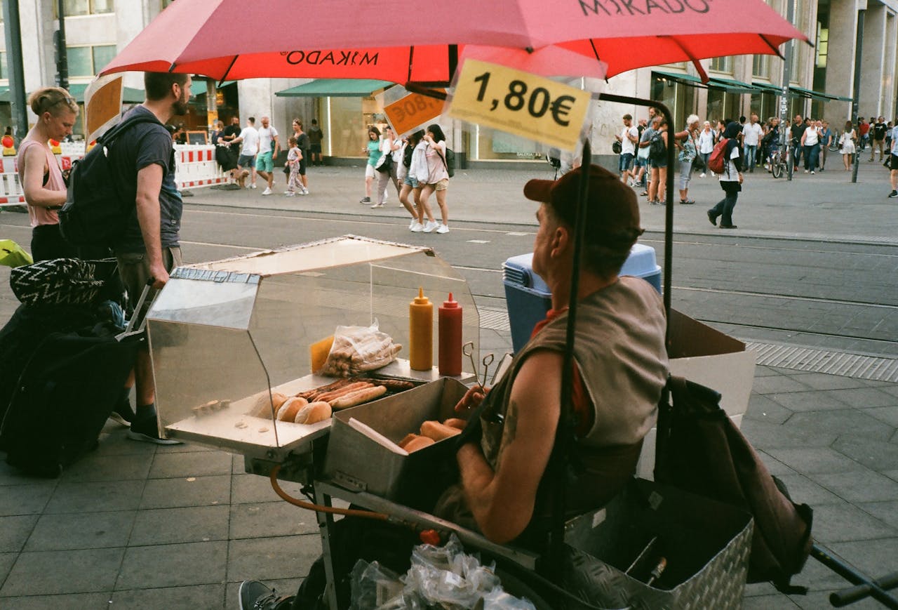 services-01 A street vendor sells hotdogs under a red umbrella in a bustling urban area.